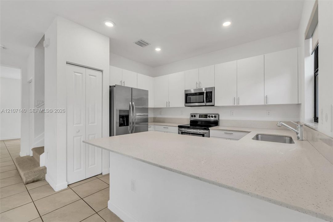 Kitchen featuring white flat-panel cabinetry, light-toned stone-finish countertops, stainless steel appliances, a peninsula with an integrated sink, and tile flooring