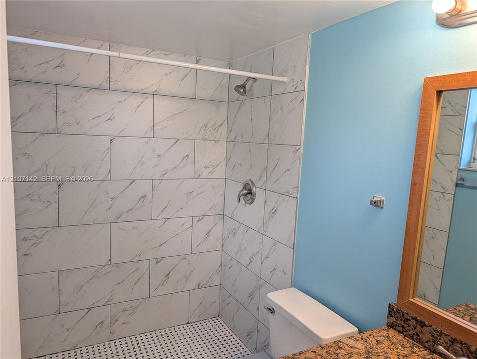 Shower enclosure featuring large-format veined wall tiles, circular mosaic floor tile, chrome shower hardware, granite-topped vanity, and a wood-framed mirror