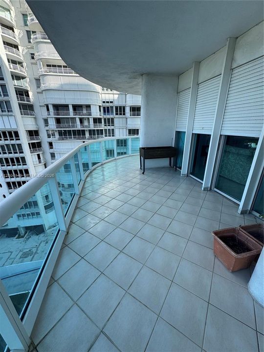 Expansive balcony featuring ceramic tile flooring, a curved glass railing with a white handrail, and integrated exterior roll-down shutters