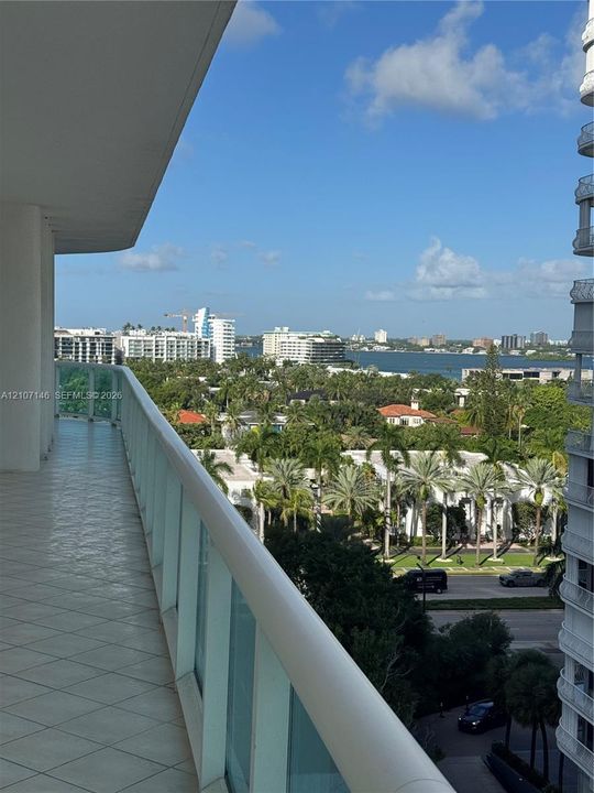 Covered balcony featuring tile flooring and a glass-paneled railing, overlooking a coastal landscape with water and urban scenery