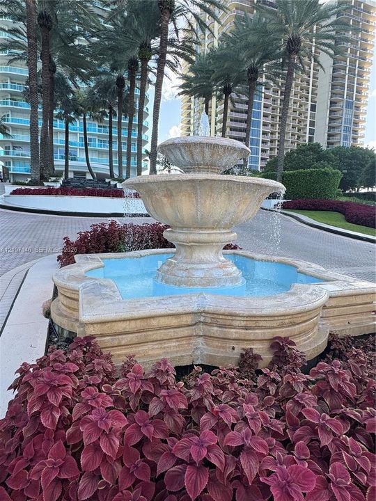 Tiered stone fountain featuring a quatrefoil basin, surrounded by manicured landscaping and mature palm trees