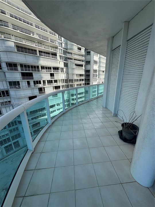Covered balcony featuring light-colored ceramic tile flooring, a curved glass railing, and integrated rolling shutters