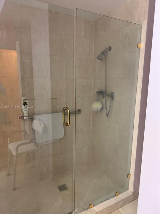 Walk-in shower featuring glass enclosure, wall-mounted shower head, horizontal grab bar, and neutral stone-finish wall and floor tiling