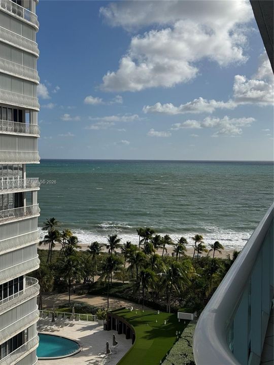 Balcony featuring a solid white railing, ocean expanse, and ground-level swimming pool with paved deck