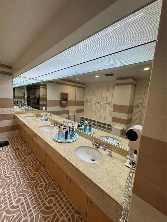 Extended vanity featuring three inset porcelain sinks, a expansive stone-surface countertop, full-width wall mirror, integrated louvered cabinetry, and patterned tile flooring