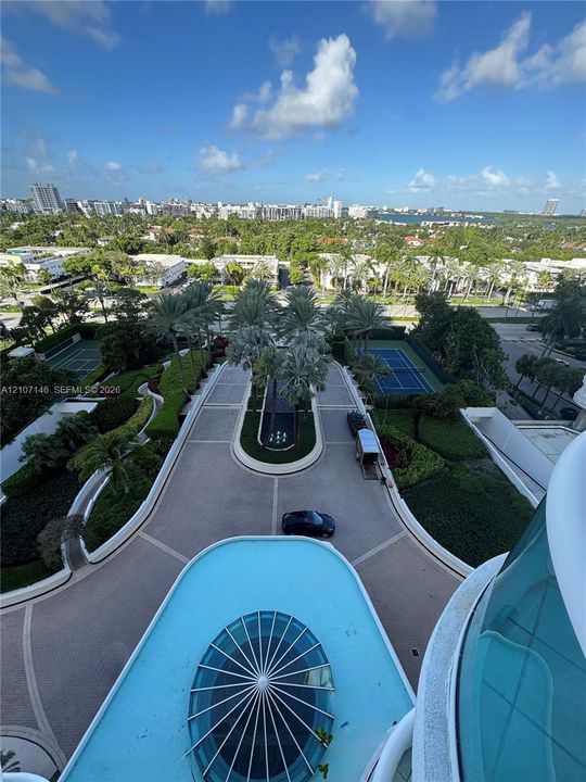 Elevated outdoor perspective featuring a circular porte-cochère, decorative water feature, manicured landscaping, and twin tennis courts