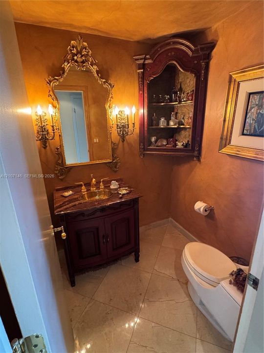 Powder room featuring a custom dark-wood vanity with a stone countertop and integrated basin, wall-mounted sconces, an ornate gold-tone framed mirror, a recessed corner display cabinet, and large-format tile flooring