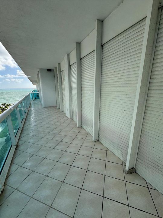 Expansive covered balcony featuring tile flooring, metal security shutters, glass-paneled railings, and coastal scenery