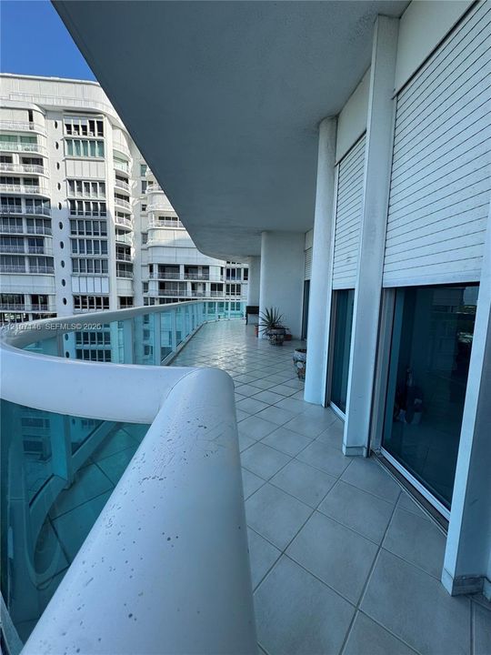 Covered wrap-around balcony featuring large-format tile flooring, a glass and metal railing system, and exterior roll-down shutters