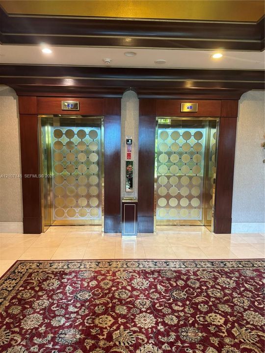 Dual elevator bank featuring custom decorative gold-toned metallic panels, dark stained wood-finish door casings, integrated crown molding, and polished stone tile flooring