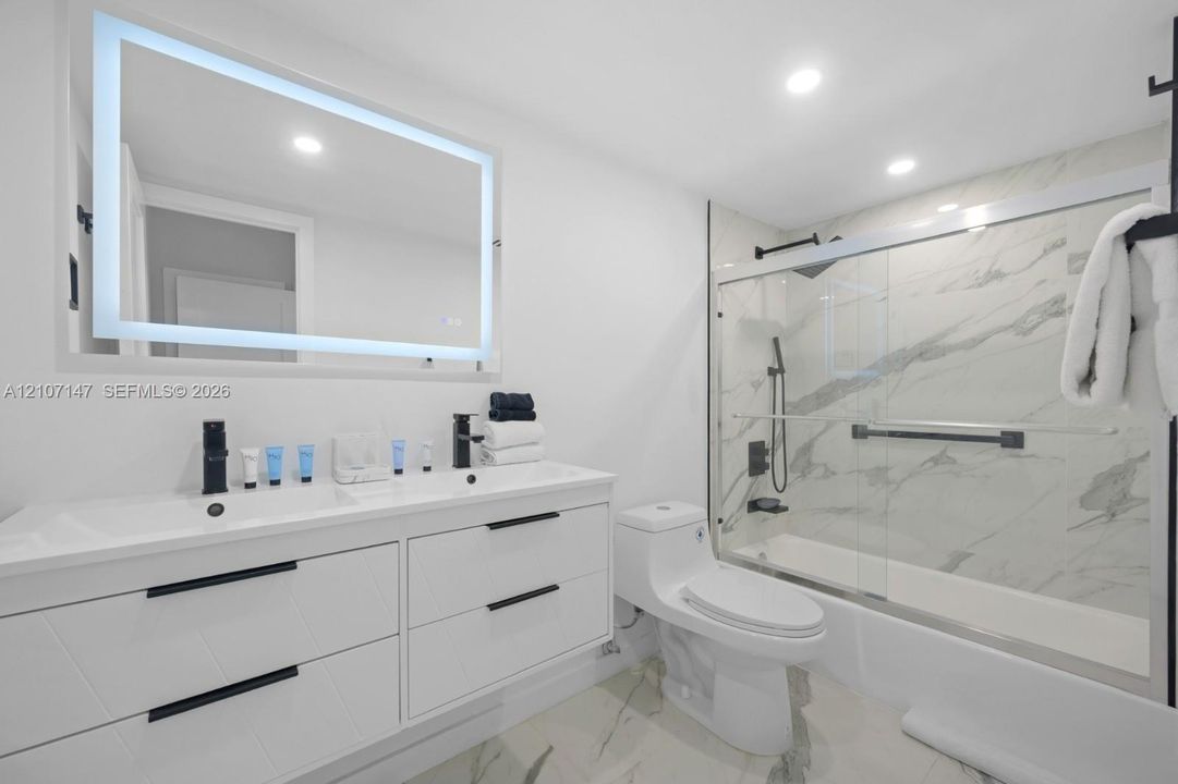 Modern bathroom featuring a double-sink vanity with matte black hardware, backlit LED mirror, recessed ceiling lighting, and a bathtub-shower combination enclosed by a glass door with large-format stone-patterned tile surround