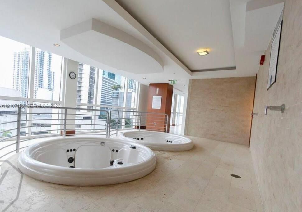 Dual jetted soaking tubs set in natural stone tile flooring, featuring floor-to-ceiling windows with a stainless steel railing and recessed ceiling lighting