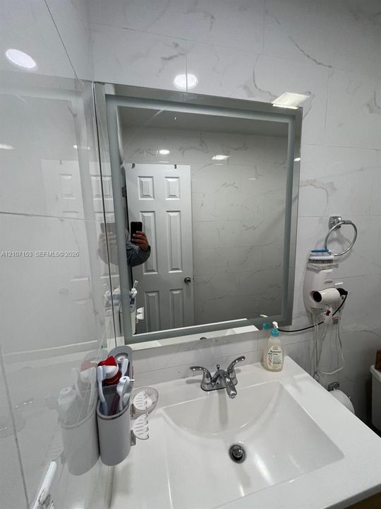 Illuminated rectangular wall mirror featuring large-format stone-patterned wall tile and a white vanity with an integrated basin and chrome dual-handle faucet