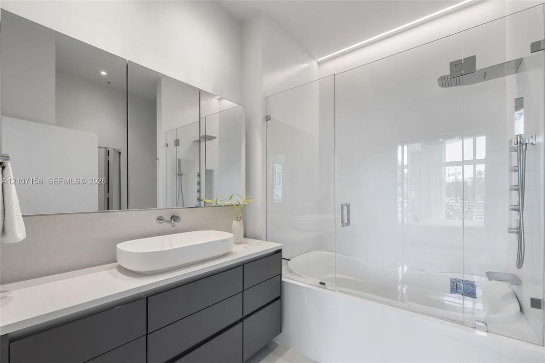 Modern vanity with charcoal cabinetry, rectangular vessel sink, white countertop, full-width mirror, and glass-enclosed shower-tub combination with chrome fixtures