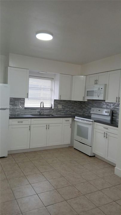 Kitchen featuring white shaker-style cabinetry, tiled backsplash, dark solid-surface countertops, tile flooring, and integrated white appliances