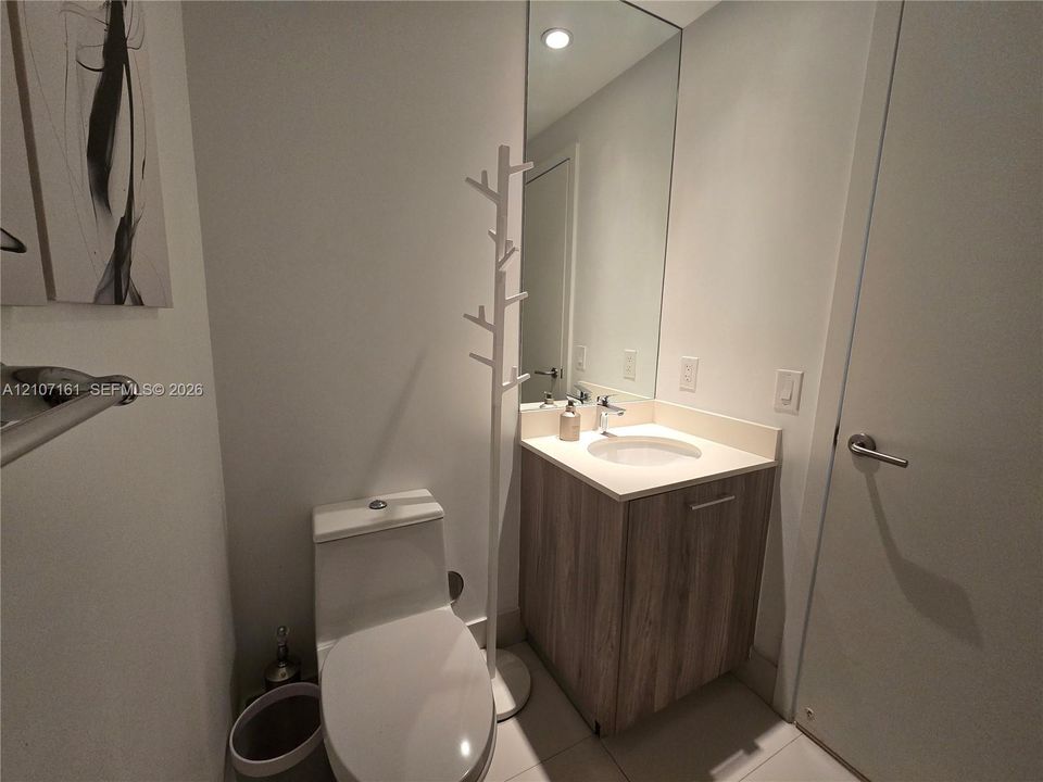 Single-flush toilet, vanity with wood-finish cabinetry, solid-surface countertop, integrated basin, and large-format wall mirror