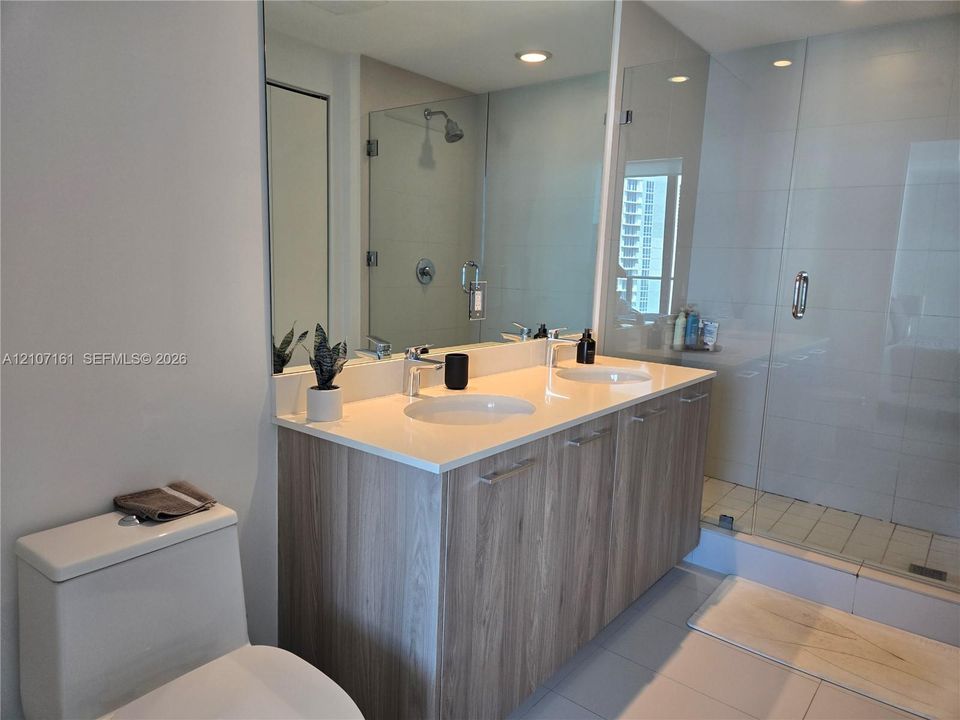 Dual-sink vanity with light wood-finish cabinetry, solid-surface countertop, full-width mirror, and a glass-enclosed shower with tiled walls