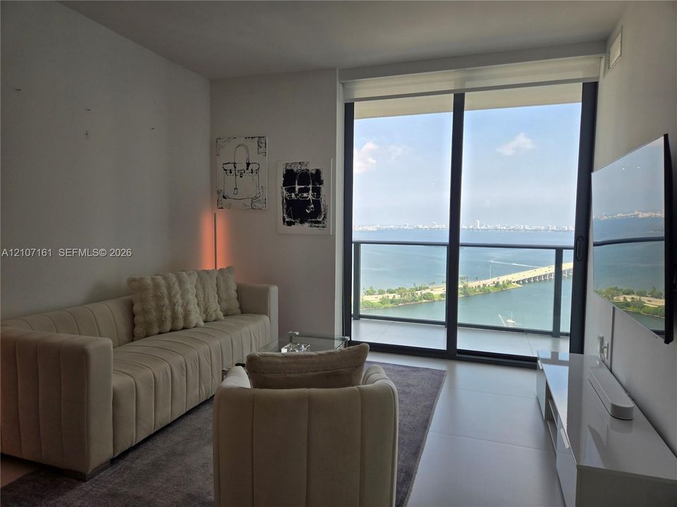 Floor-to-ceiling glass sliding doors opening to a balcony with expansive water and causeway scenery, complemented by large-format tile flooring