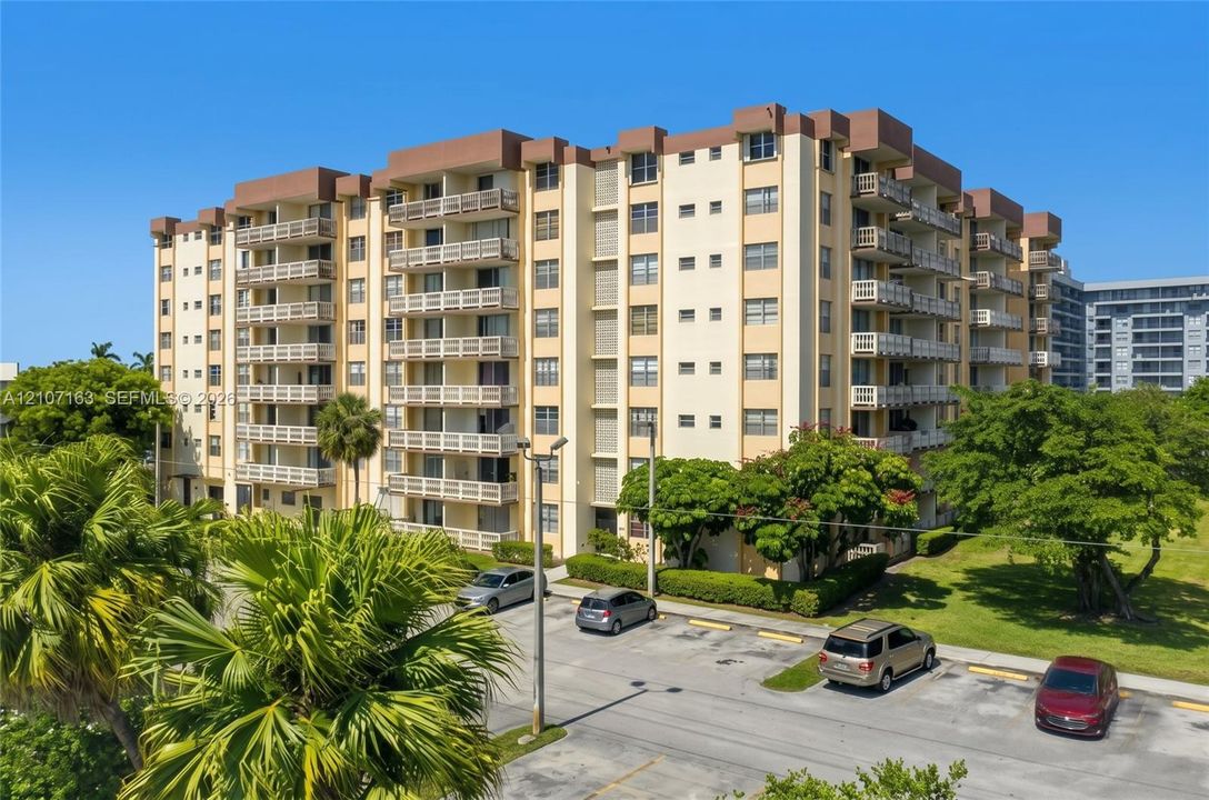 Multi-story residential building featuring private balconies, masonry exterior, concrete parking surface, and established landscaping