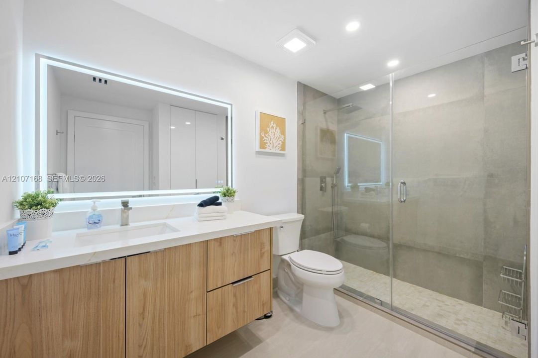 Bathroom featuring a wood-finish vanity with a stone countertop, an integrated rectangular sink, a frameless glass-enclosed shower, and a large backlit mirror