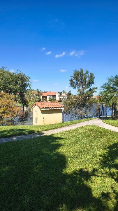 Expansive lawn area featuring paved walkways, a utility structure with a red tile roof, and waterfront perimeter fencing