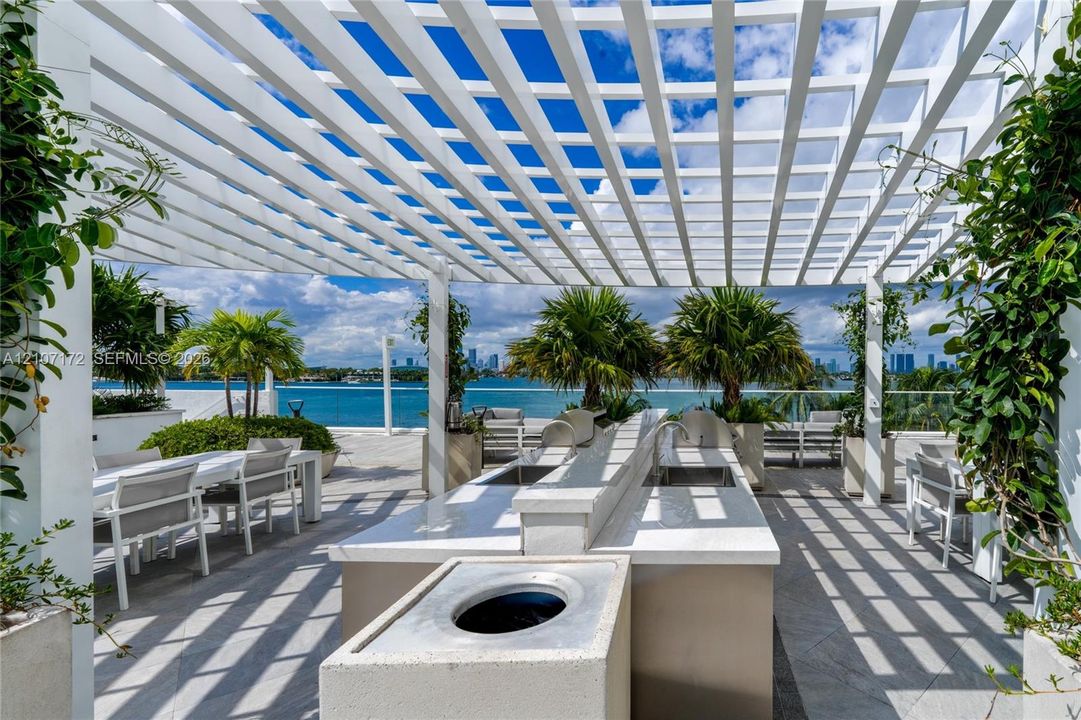Outdoor terrace featuring a white louvered pergola, built-in kitchen island with stone countertops and stainless steel sink, tile flooring, and glass balcony railings