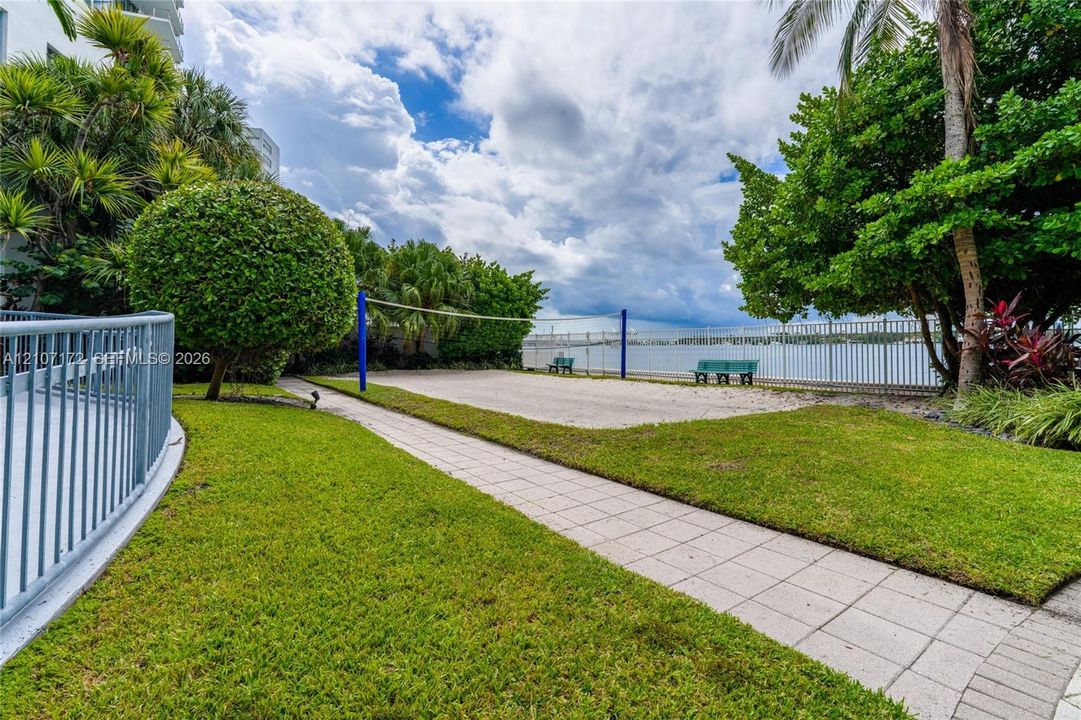 Sand volleyball court featuring a net, bordering a paved stone walkway, manicured lawn, and water frontage