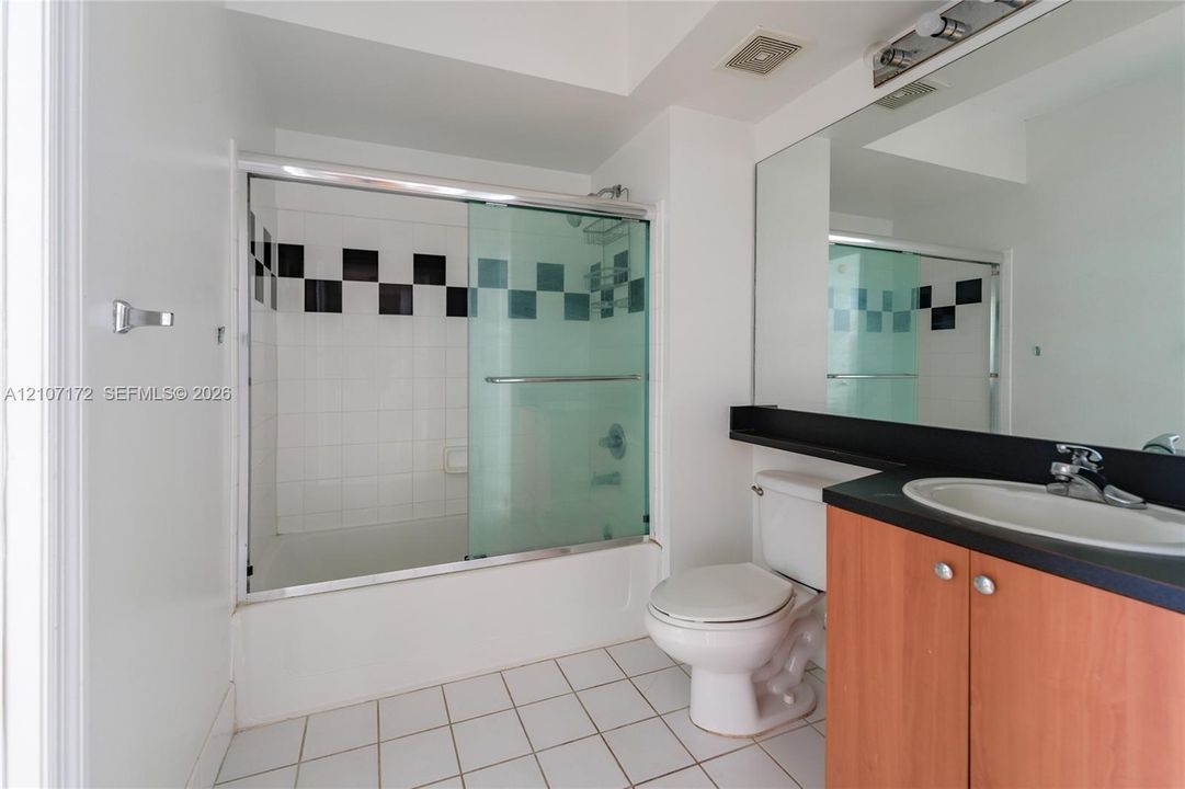 Bathroom featuring a vanity with wood-finish cabinetry, a black countertop with an integrated oval sink, a wall-to-wall mirror, and a shower-tub combination with sliding glass doors and decorative tile inlay