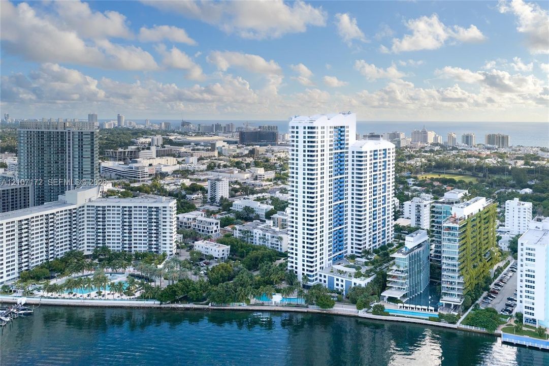 Aerial perspective of a high-rise residential complex featuring waterfront positioning, multiple outdoor swimming pools, landscaped grounds, and proximity to coastal waters