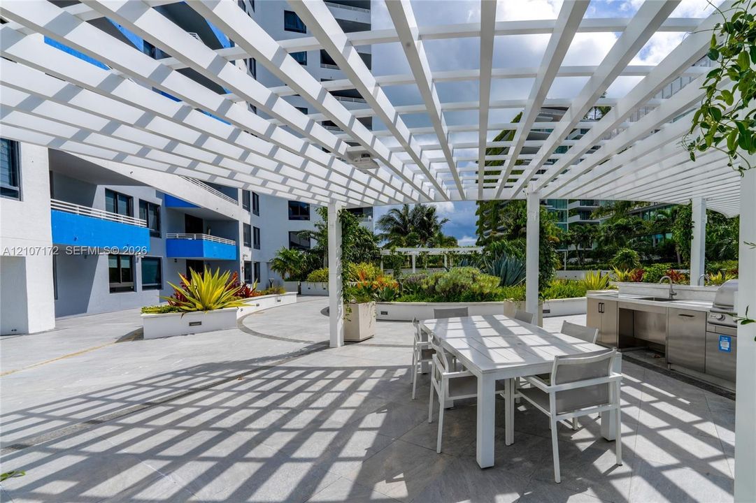 Outdoor terrace featuring a white overhead trellis, built-in stainless steel kitchen station, stone-surfaced flooring, and perimeter planters