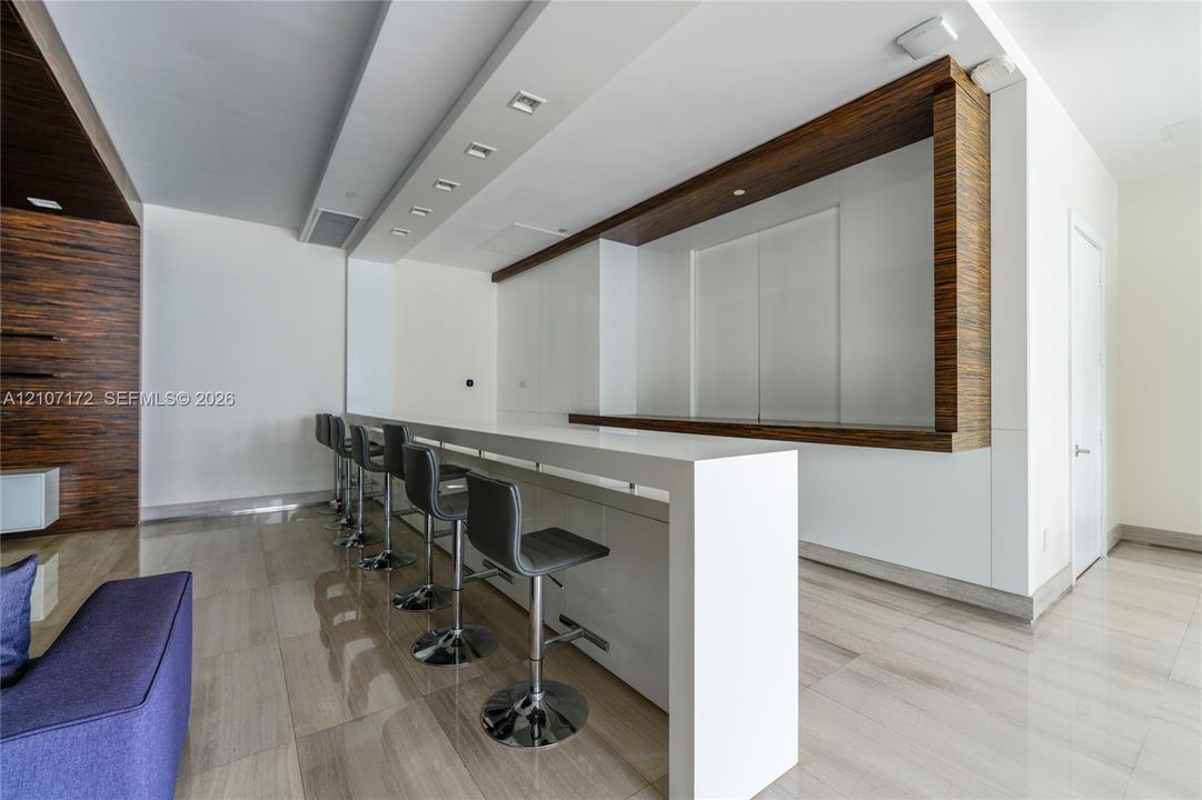 Custom built-in bar featuring a sleek waterfall edge countertop, integrated wood-finish cabinetry, recessed ceiling lighting, and polished stone flooring