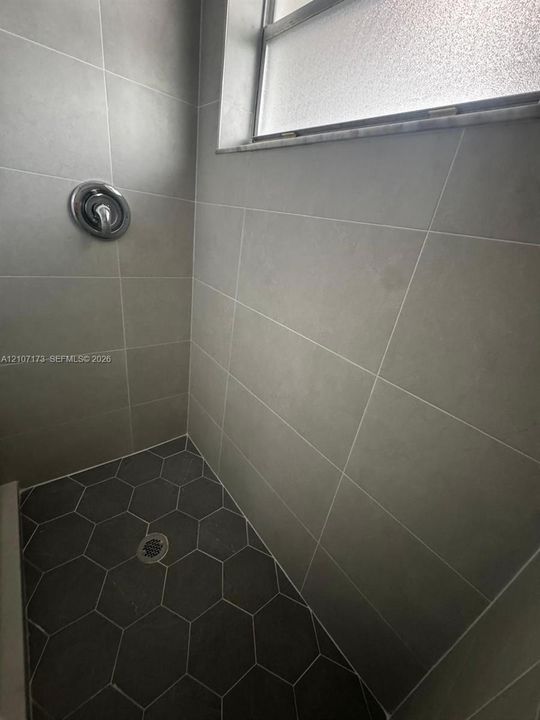 Shower enclosure featuring large-format grey wall tile, dark charcoal hexagonal floor tile, a polished chrome shower fixture, and an obscured-glass window