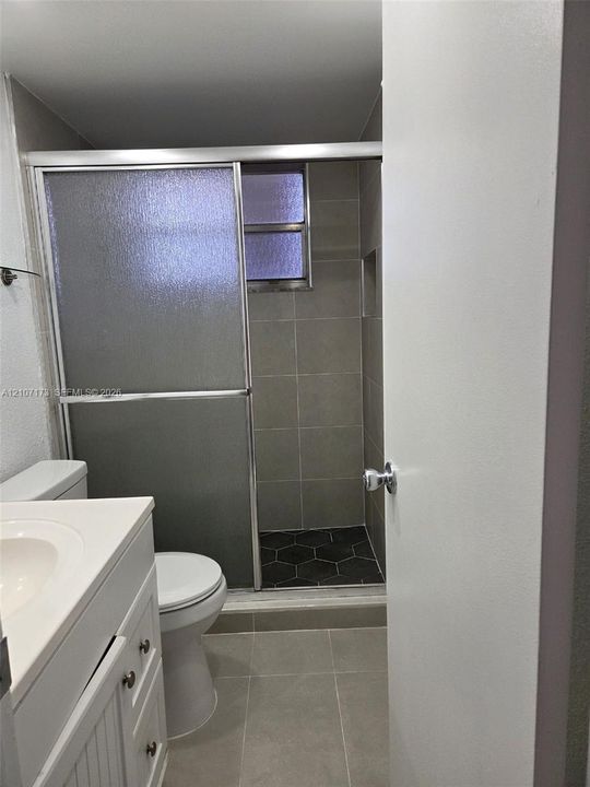 Bathroom featuring a white vanity with an integrated sink, walk-in shower with sliding glass doors, neutral large-format tile flooring, and contrasting black hexagonal floor tiling within the shower
