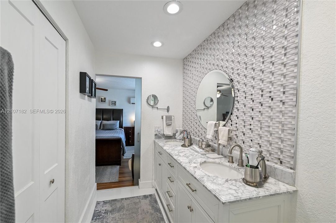 Dual-vanity cabinetry with stone-finish countertops, mosaic-tiled accent wall, circular wall-mounted mirror, and recessed lighting