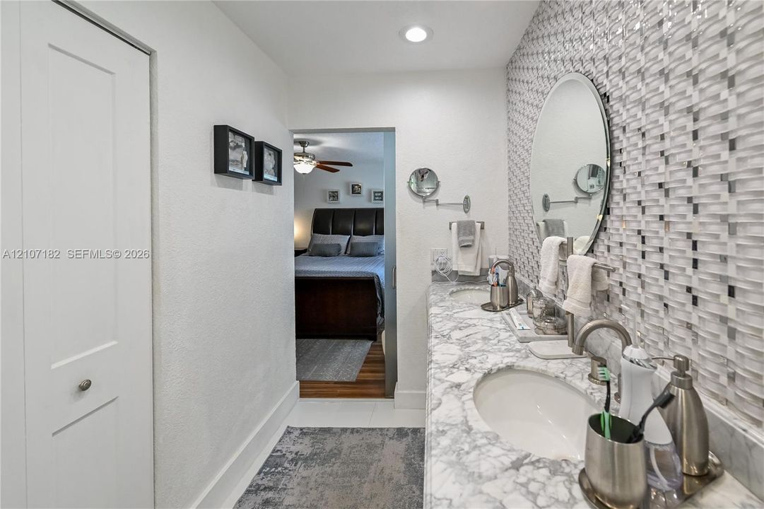 Dual-sink vanity with patterned stone countertop, custom mosaic tile backsplash, recessed lighting, and oval wall-mounted mirrors
