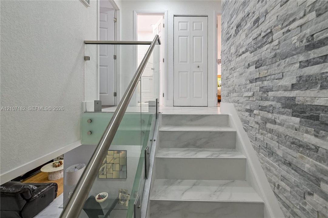 Staircase featuring white stone-finish treads, a stacked-stone accent wall, and a glass panel railing with a metallic handrail