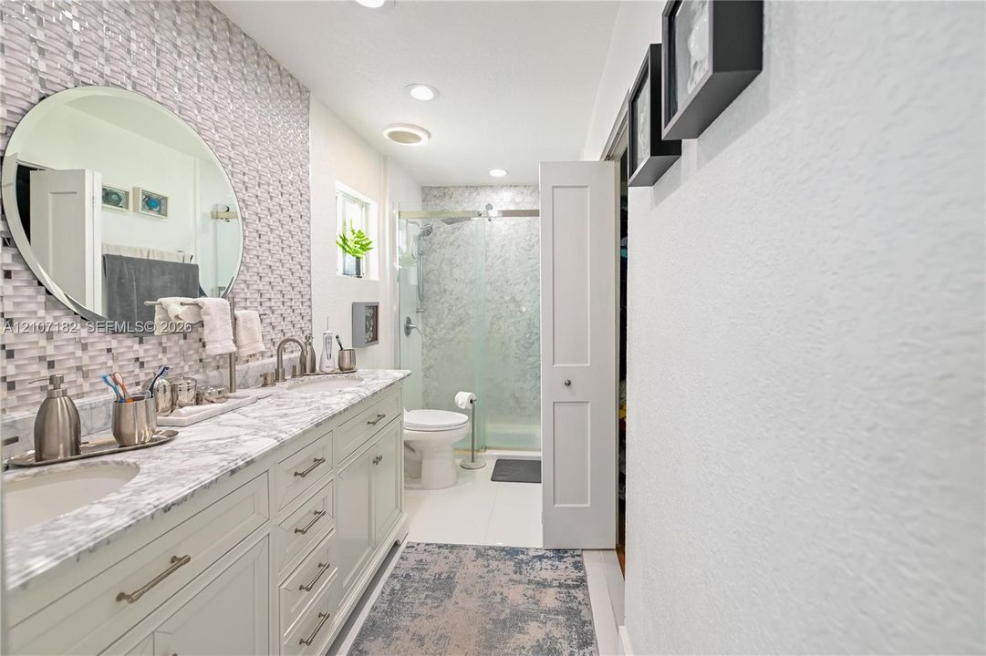 Bathroom featuring a vanity with stone-finish countertop, dual under-mount sinks, a circular wall-mounted mirror, decorative tiled backsplash, and a glass-enclosed shower