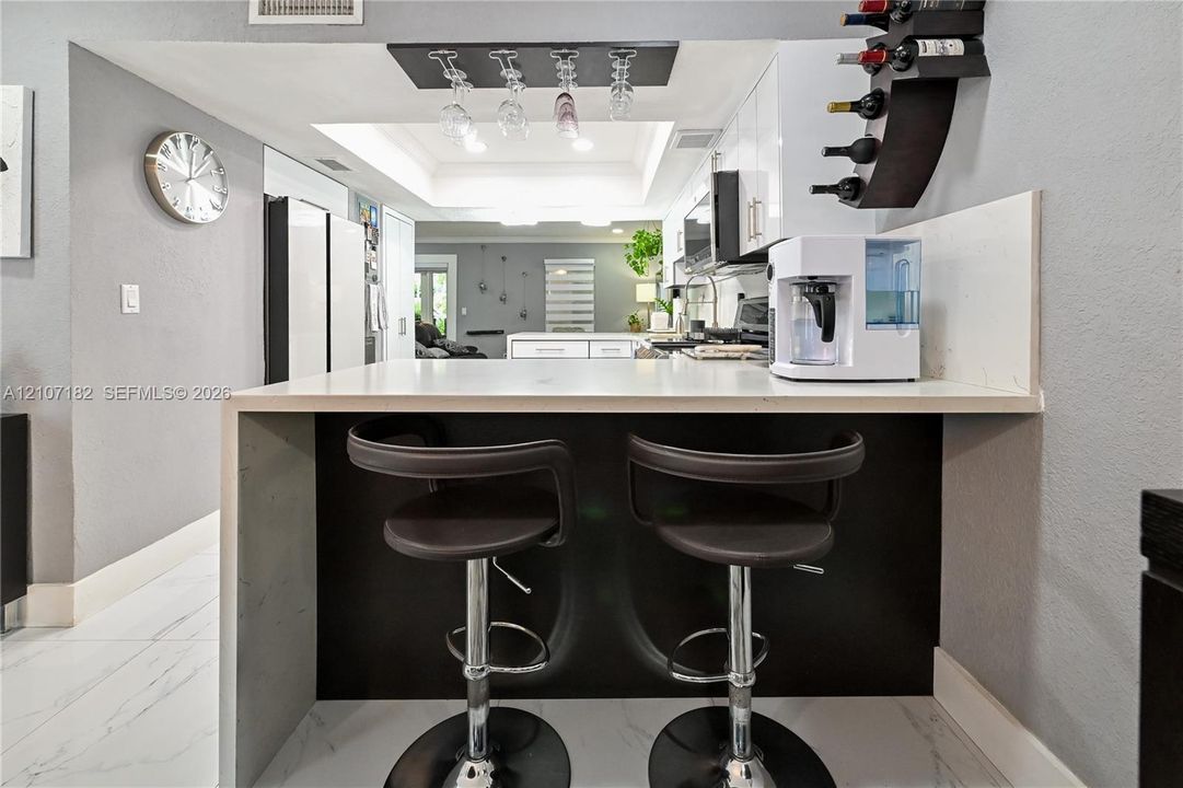 Waterfall peninsula featuring a solid-surface countertop and contrasting dark cabinetry, overhead wine glass rack, tray ceiling, and white floor tiling with veining