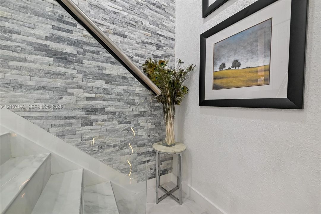 Staircase featuring stacked stone accent wall, glass panel railing, and white stone-finish treads