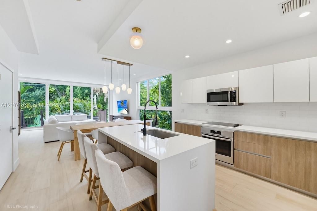 Modern kitchen featuring a central island with a quartz countertop, white flat-panel upper cabinetry, wood-finish lower cabinetry, integrated stainless steel appliances, and large-format windows