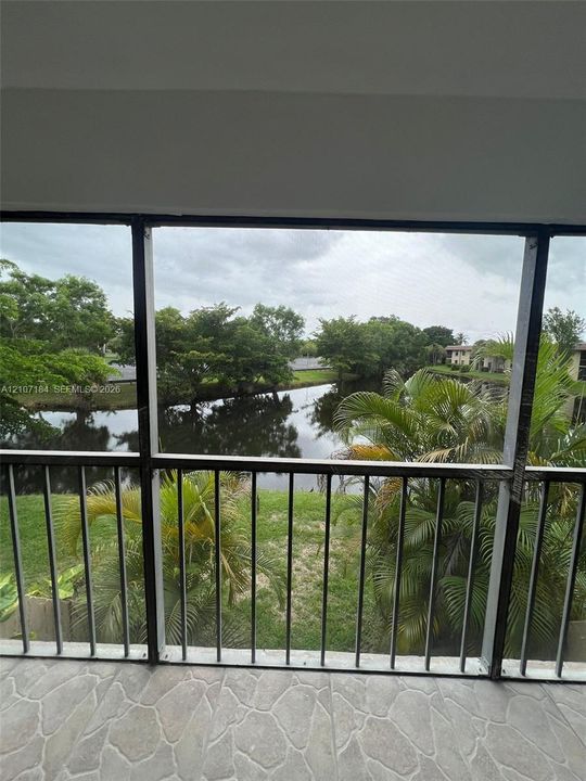 Screened-in balcony featuring stone-finish tile flooring, metal railing, and direct exposure to a water feature with surrounding mature landscaping