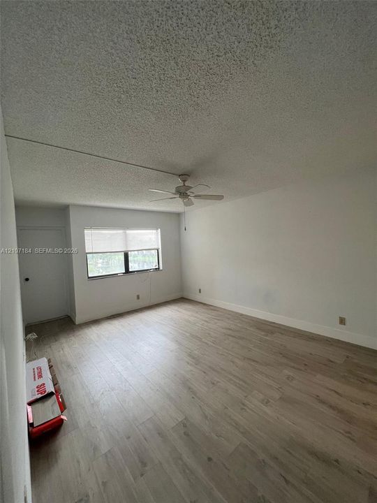 Room featuring wood-finish flooring, a textured ceiling, a central ceiling fan with integrated lighting, and a window with a roller shade