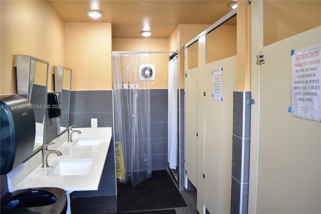 Nearby pool cabana facility featuring a dual-sink vanity with white countertop, integrated basin, and metal faucets, finished with gray tile wainscoting and multiple privacy partitions