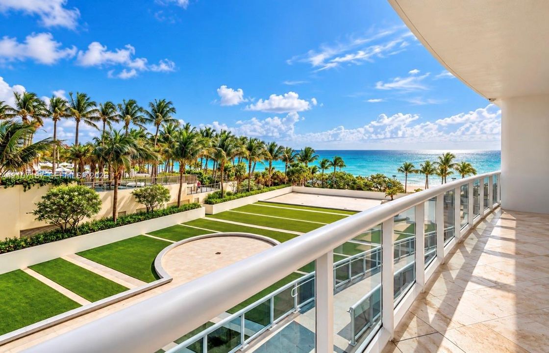 Private balcony featuring stone tile flooring and a glass-paneled railing overlooking a landscaped courtyard with artificial turf, stone accents, and a coastal backdrop