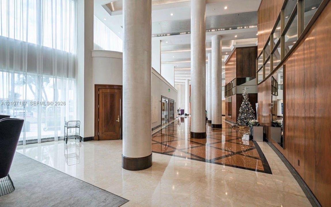Grand lobby featuring floor-to-ceiling windows, soaring ceilings with recessed lighting, polished stone flooring with geometric tile inlay, full-height architectural columns, and wood-paneled wall accents