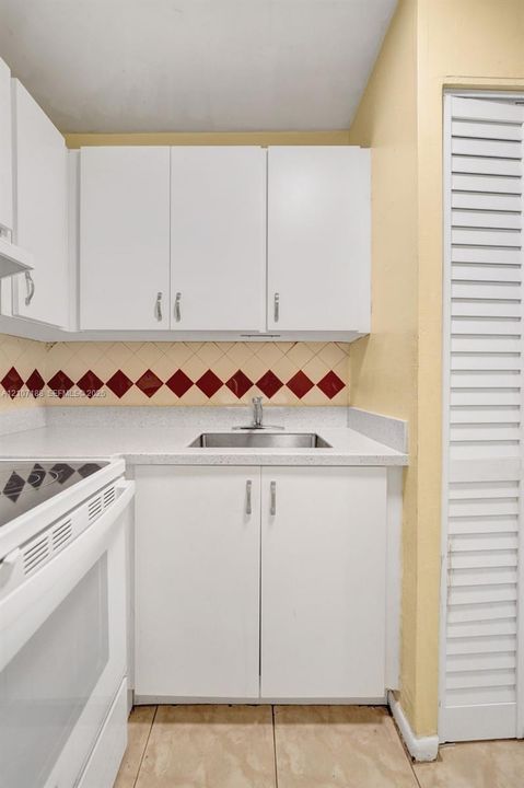 Kitchenette featuring white cabinetry, a neutral solid-surface countertop, stainless steel sink with a chrome faucet, diamond-patterned accent tile backsplash, and louvered closet doors