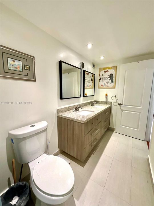Double vanity featuring light wood-finish cabinetry, stone-composite countertop with integrated dual sinks, two rectangular mirrors with black frames, and tile flooring