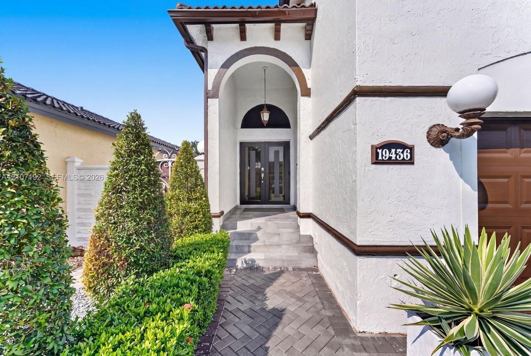 Stucco exterior featuring an arched entryway with a hanging pendant light, dual-pane glass front doors, herringbone-pattern brick paver walkway, and stone steps