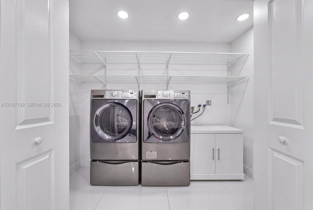 Laundry area featuring large-format tile flooring, recessed lighting, white base cabinetry with dual doors, tiered wire shelving, and double-door entry