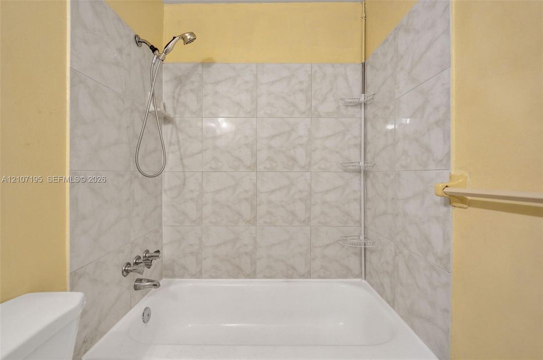 Shower and tub combination featuring light-toned tile surround, handheld shower head, chrome-finish faucet fixtures, and integrated wall-mounted shelving